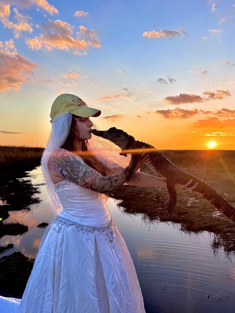Woman in a wedding dress and veil holding a small alligator at sunset